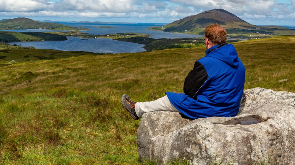 hiker resting on a rock admiring the view in the Irish countryside with lakes, mountains and hills in the background, day with a blue sky and white clouds in Letterfrack